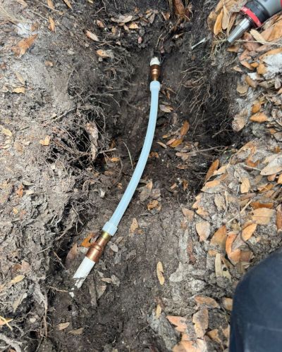 plumbing installation of a water line in the ground with copper fittings and a clear plastic hose visible among dirt and leaves