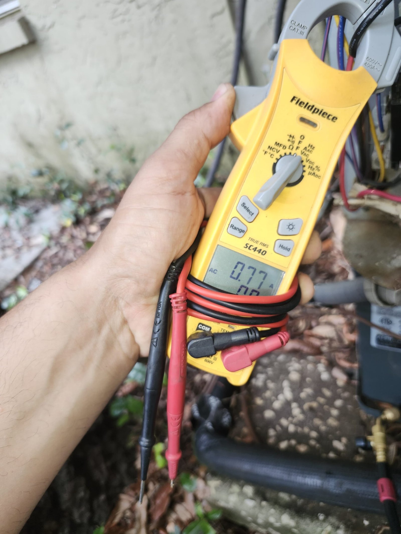 Hand holding multimeter with visible readings near AC unit in Lutz, FL 33559. Diagnostic process.