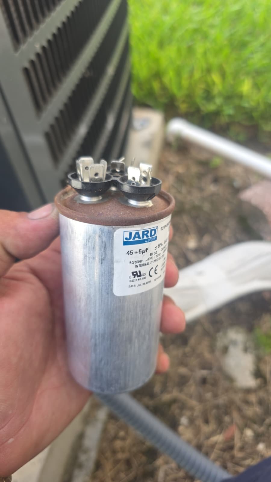 Hand holding JARD capacitor in front of outdoor AC unit, Tampa, FL 33618.