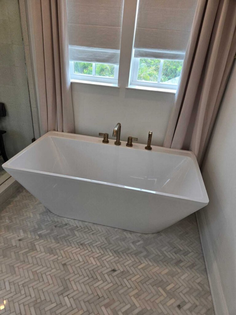 a modern bathtub installed with bronze fixtures in a well-lit bathroom featuring herringbone patterned flooring and window coverings