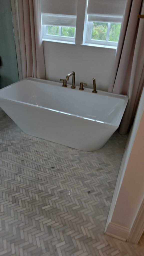a modern bathtub installed with gold faucets on a herringbone marble floor in a well-lit bathroom
