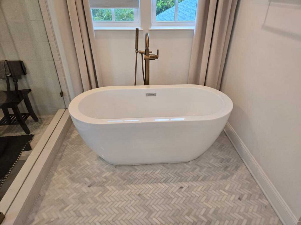 a modern freestanding bathtub situated on herringbone patterned tile in a well-lit bathroom with natural light