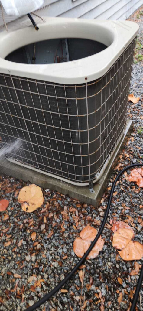 cleaning an air conditioning unit with a hose on a gravel surface surrounded by fallen leaves