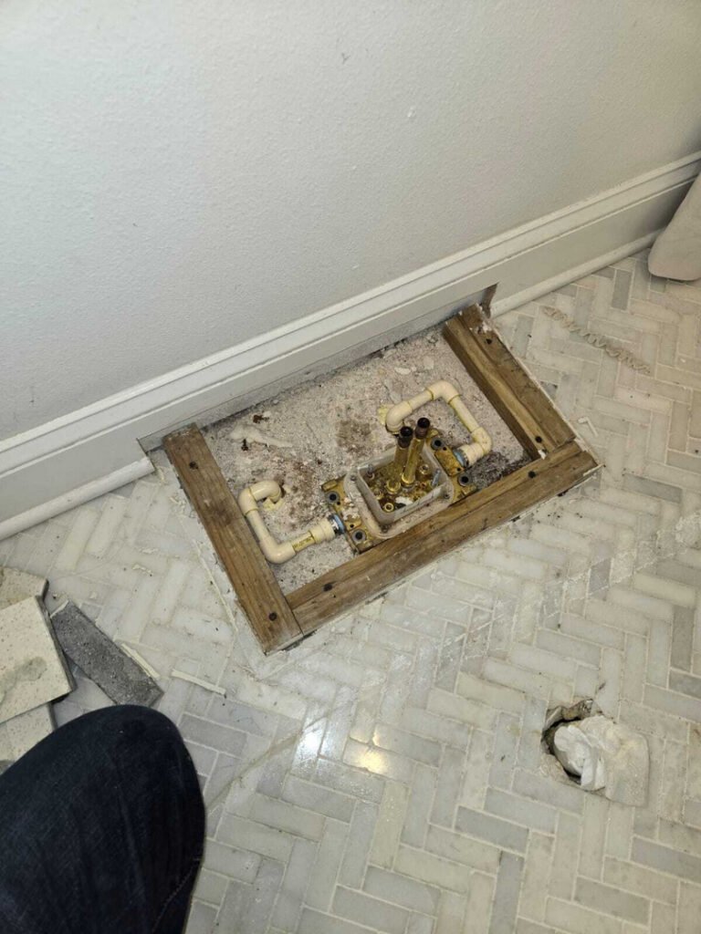 exposed plumbing under a floor with a wooden frame and some debris around it including tiles and a small hole in the floor