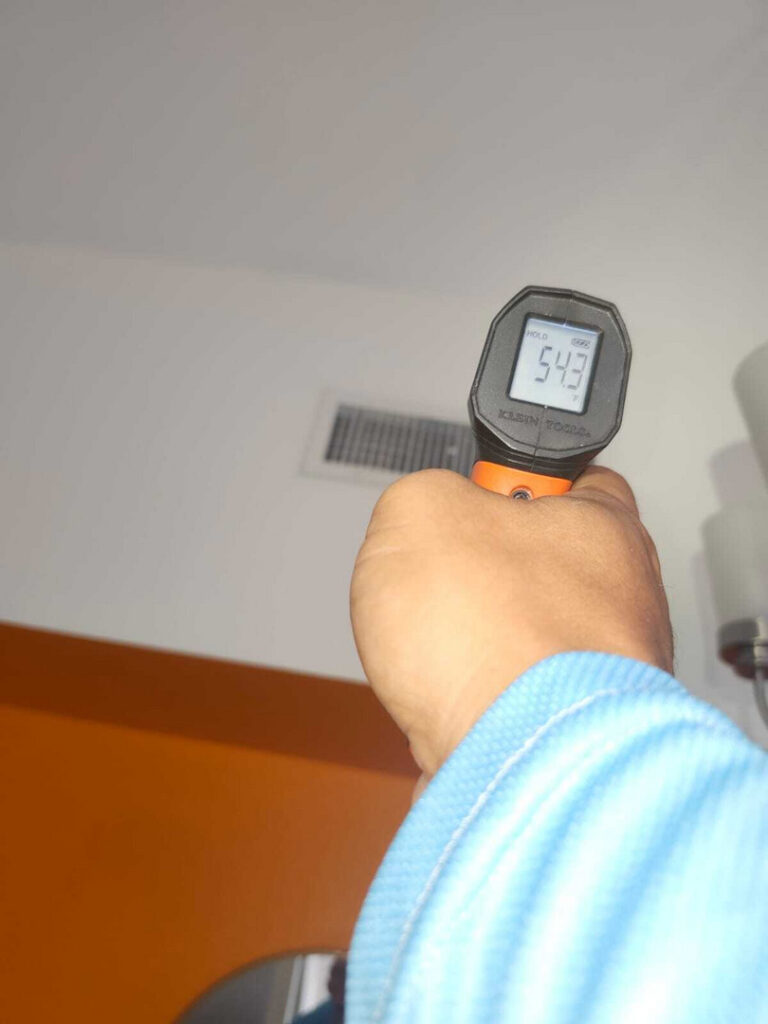 holding a digital thermometer measuring temperature in a room with visible air vent and orange wall behind