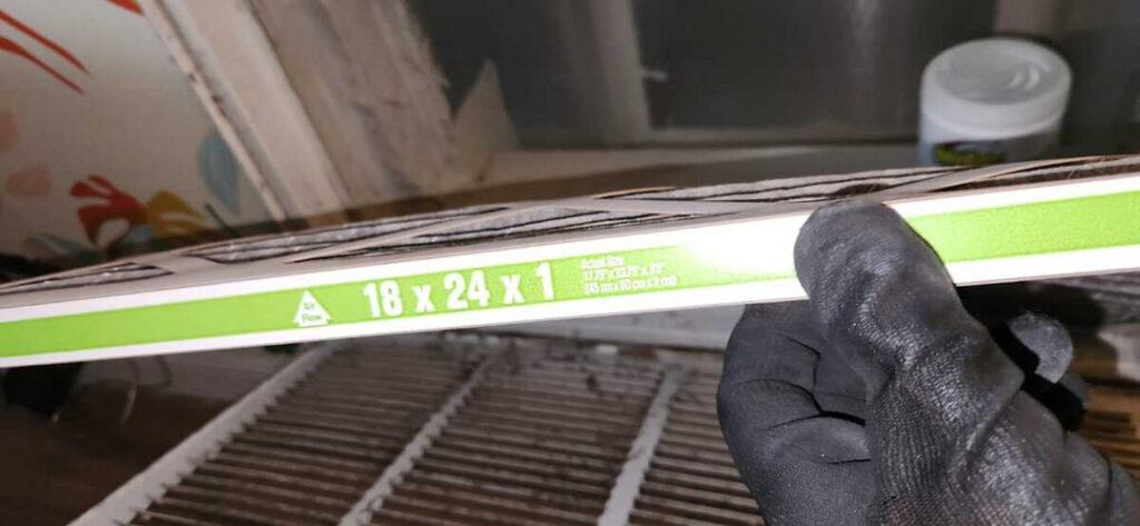 holding a new air filter measuring eighteen by twenty-four by one inches in a gloved hand near a vent