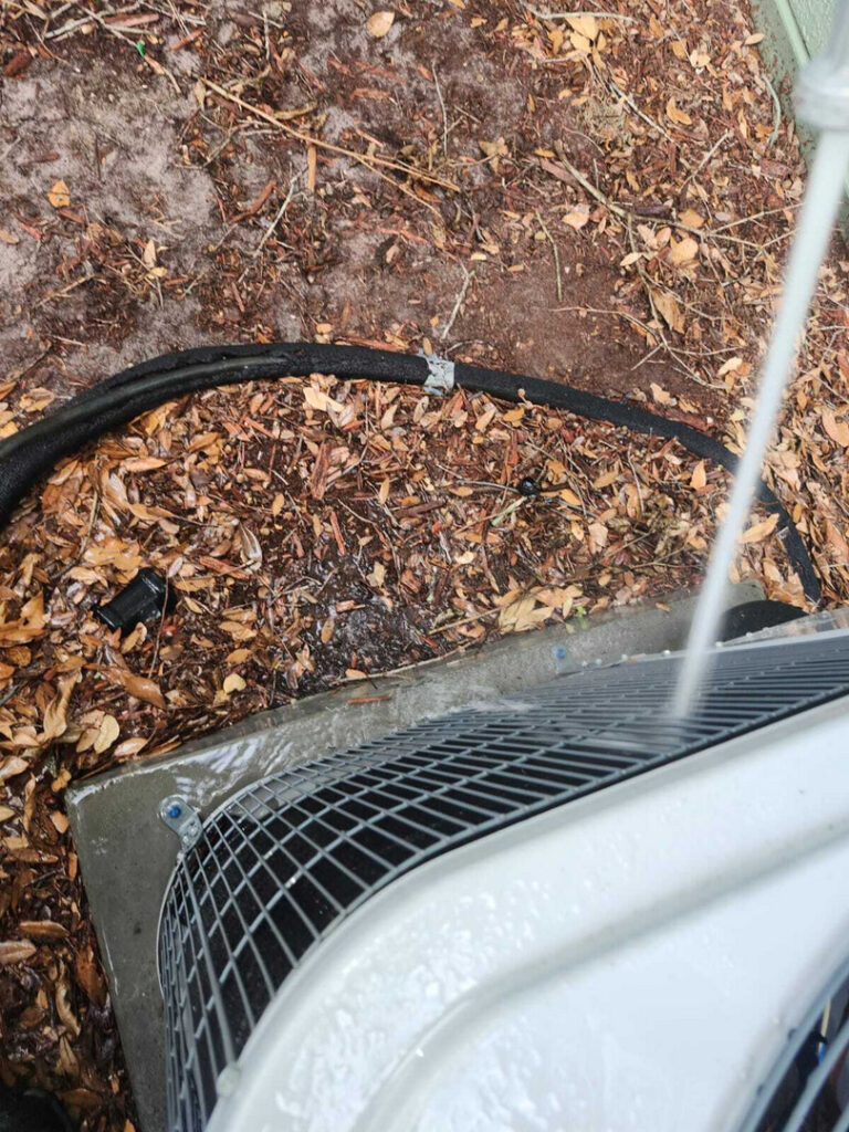 home therapist cooling heating and plumbing cleaning an air conditioning unit with water while surrounded by leaves and dirt