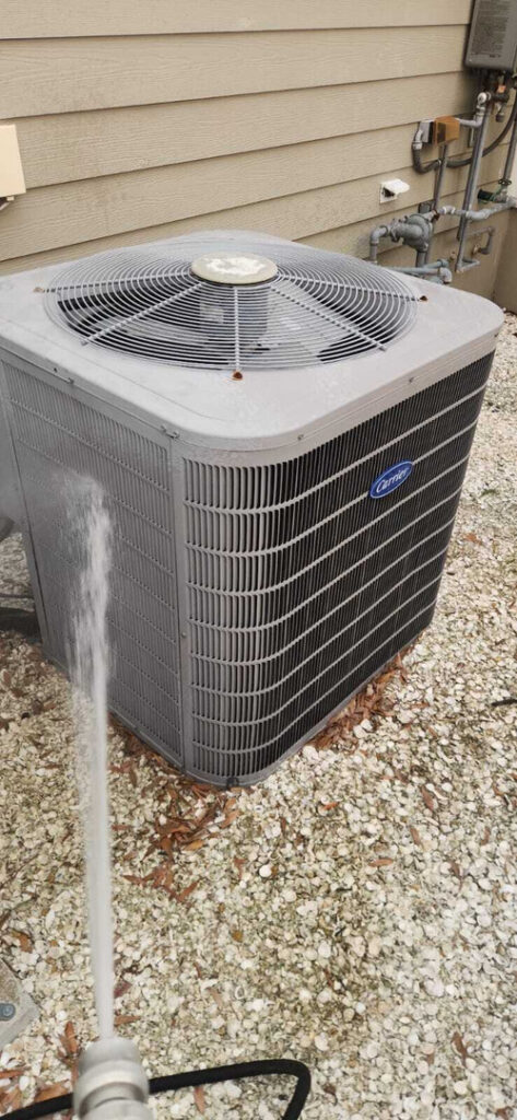 home therapist cooling heating and plumbing cleaning an outdoor air conditioning unit with a pressure washer near gravel ground