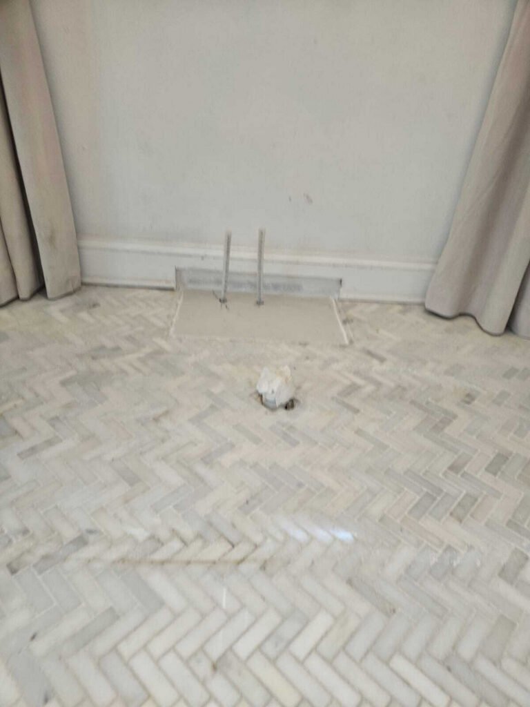 marble flooring with a floor access panel and exposed pipes near a wall with curtains and herringbone pattern