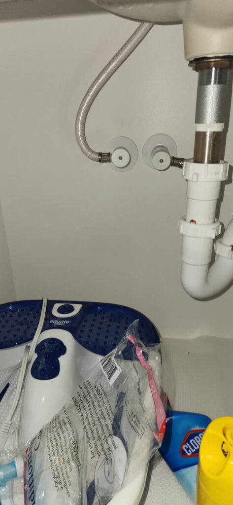 plumbing work under a sink with pipes and hoses visible alongside cleaning supplies and a foot spa