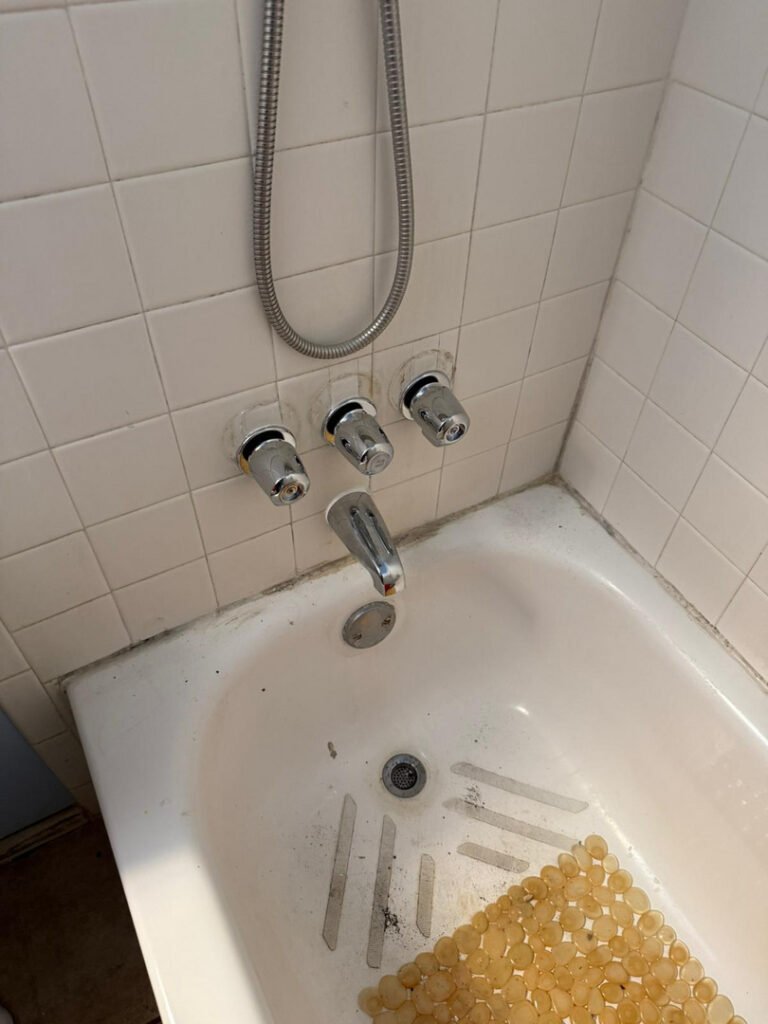 three faucet handles and showerhead visible in a bathtub with dirt around the drain and some water spots
