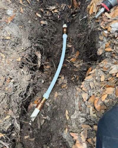 plumbing installation of a water line in the ground with copper fittings and a clear plastic hose visible among dirt and leaves
